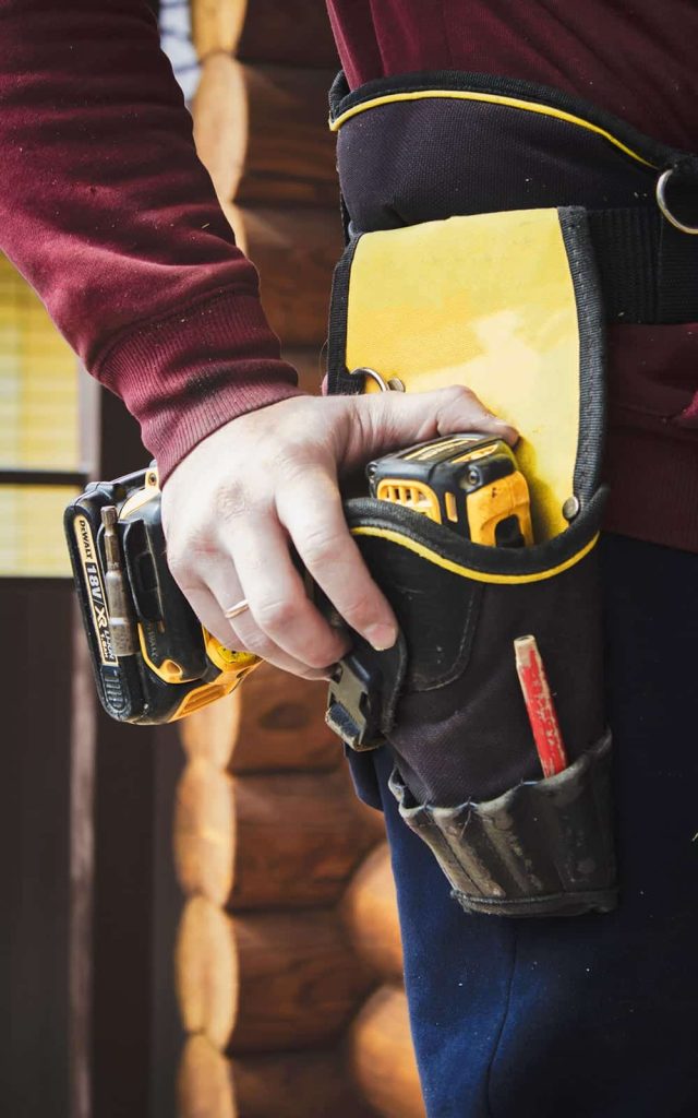 A person wearing a tool belt with a drill and a red-handled tool is dressed in a maroon long-sleeve shirt and dark pants, standing in front of a log cabin wall, possibly involved in tasks such as scale calibration.
